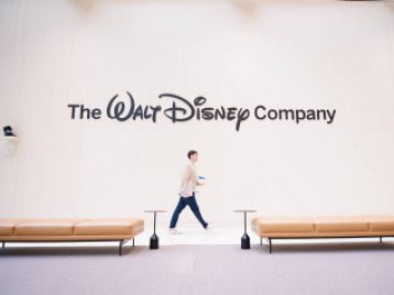 Wide shot of the Walt Disney Company logo on a wall, with two chaise lounges on either side and a Disney employee walking past.